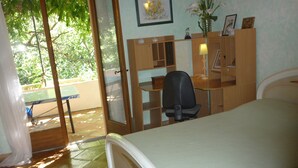 A bedroom with a bed, a desk, and a chair. There is a window with a view of trees and a door leading to an outdoor area.