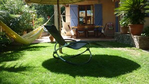 A hammock, a chair, and a table under a thatched umbrella in a garden area.