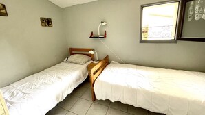 A small, simple room with two single beds, a desk with a lamp, and a window with a decorative iron grille.