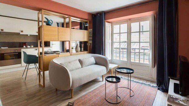 A modern living room with a sofa, a coffee table, and a bookshelf. There is a window with curtains and a view of buildings outside.