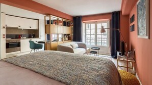 A compact hotel room with a bed, a desk, a kitchenette, and a balcony.