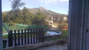 Une vue à travers une fenêtre donnant sur une cour arrière avec une clôture en bois, une petite piscine et une montagne en arrière-plan.