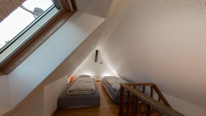 Une petite pièce étroite avec un plafond en pente et un escalier en bois. On y trouve deux lits avec du linge de lit blanc et une petite table avec une lampe.