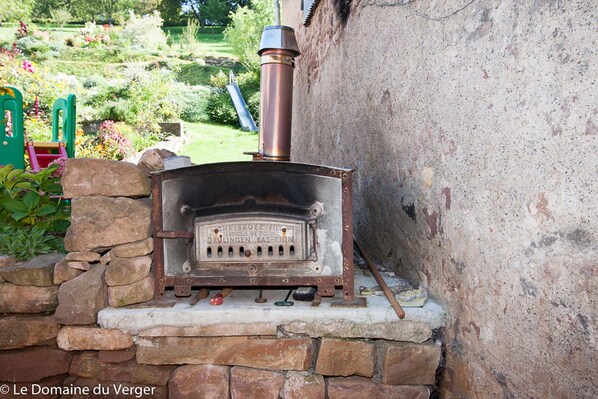 A rustic outdoor wood stove with a chimney, set against a stone wall in a garden.