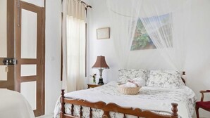 A bedroom with a wooden bed, a nightstand with a lamp, a wicker basket, and a mosquito net. - Lefkara View (Pano Lefkara)