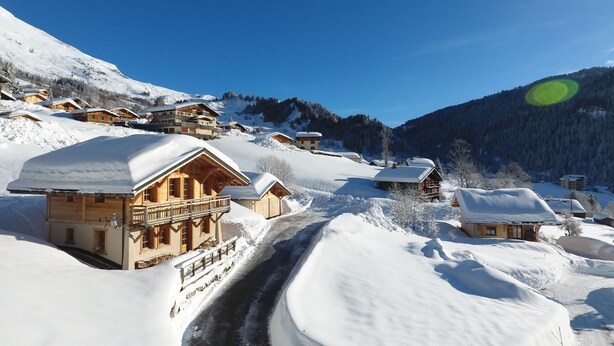 A snow-covered village with wooden chalets, a clear sky, and a mountain backdrop.