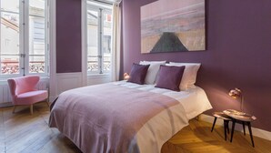 A bedroom with a bed, a pink chair, a bedside table with a lamp, and a large painting on the wall. - Confort - Furnished apartment - Downtown Lyon (Lyon)