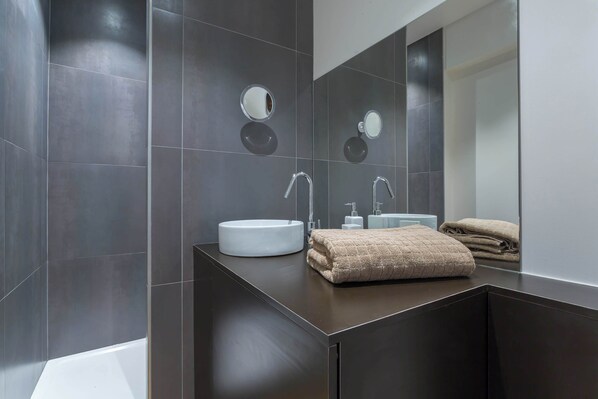 A modern bathroom with dark tiles, a white sink, a mirror, and a towel. - Confort - Furnished apartment - Downtown Lyon (Lyon)