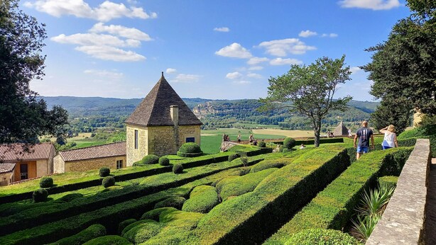 Un bâtiment historique en pierre, surmonté d’un toit conique, entouré de haies bien entretenues et offrant une vue sur des collines verdoyantes à l’horizon.