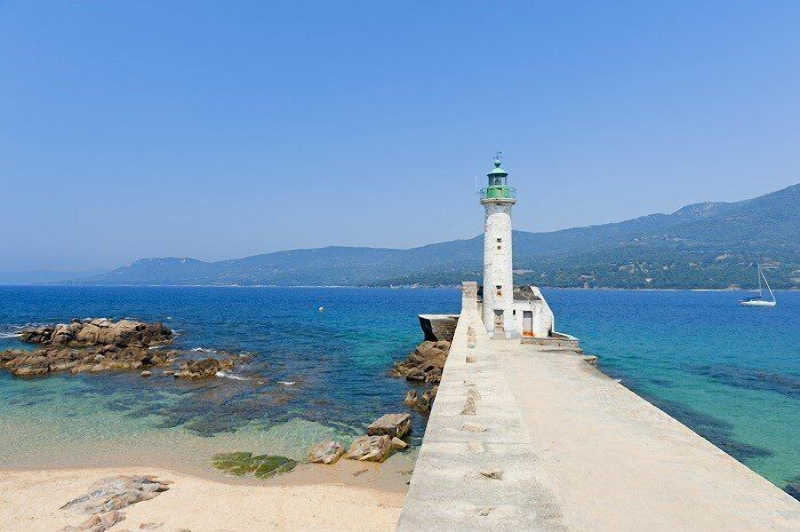 A lighthouse on a pier extending into a clear blue sea with rocky outcrops and a distant mountain range.