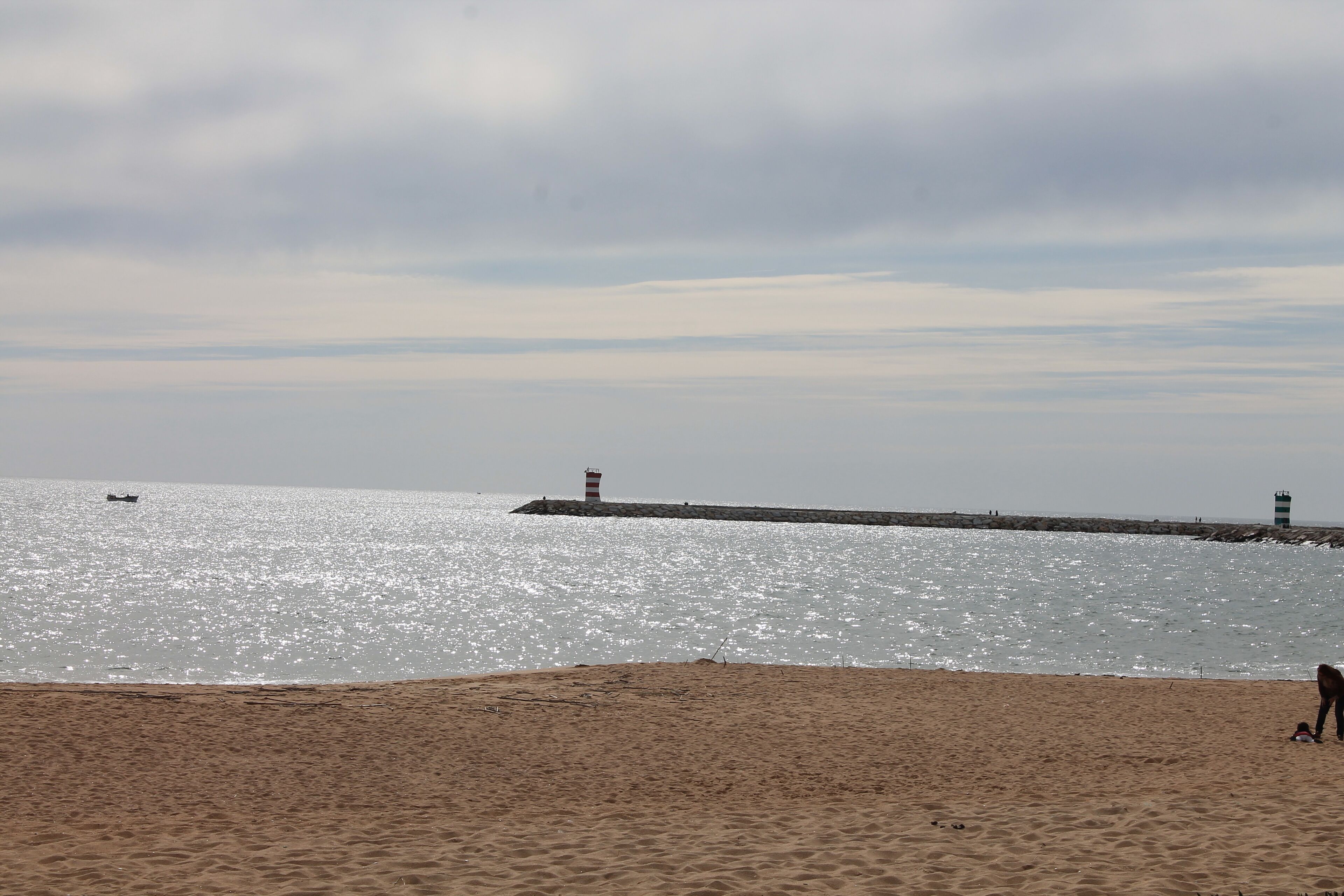 A beach with a pier extending into the water, a boat in the distance, and a person walking on the sand.