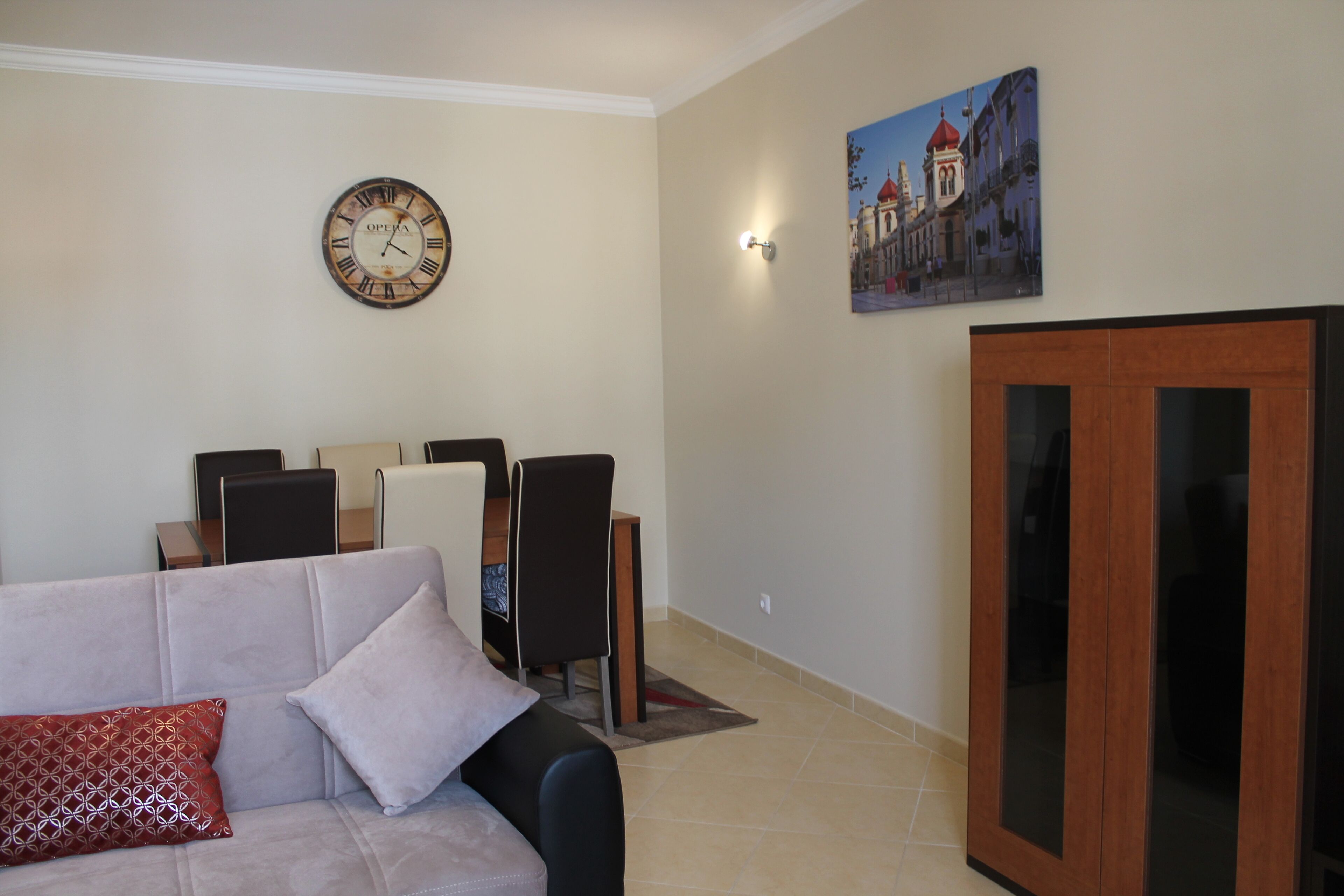 A living room with a sofa, a dining table with chairs, a wooden cabinet, a wall clock, and a painting.