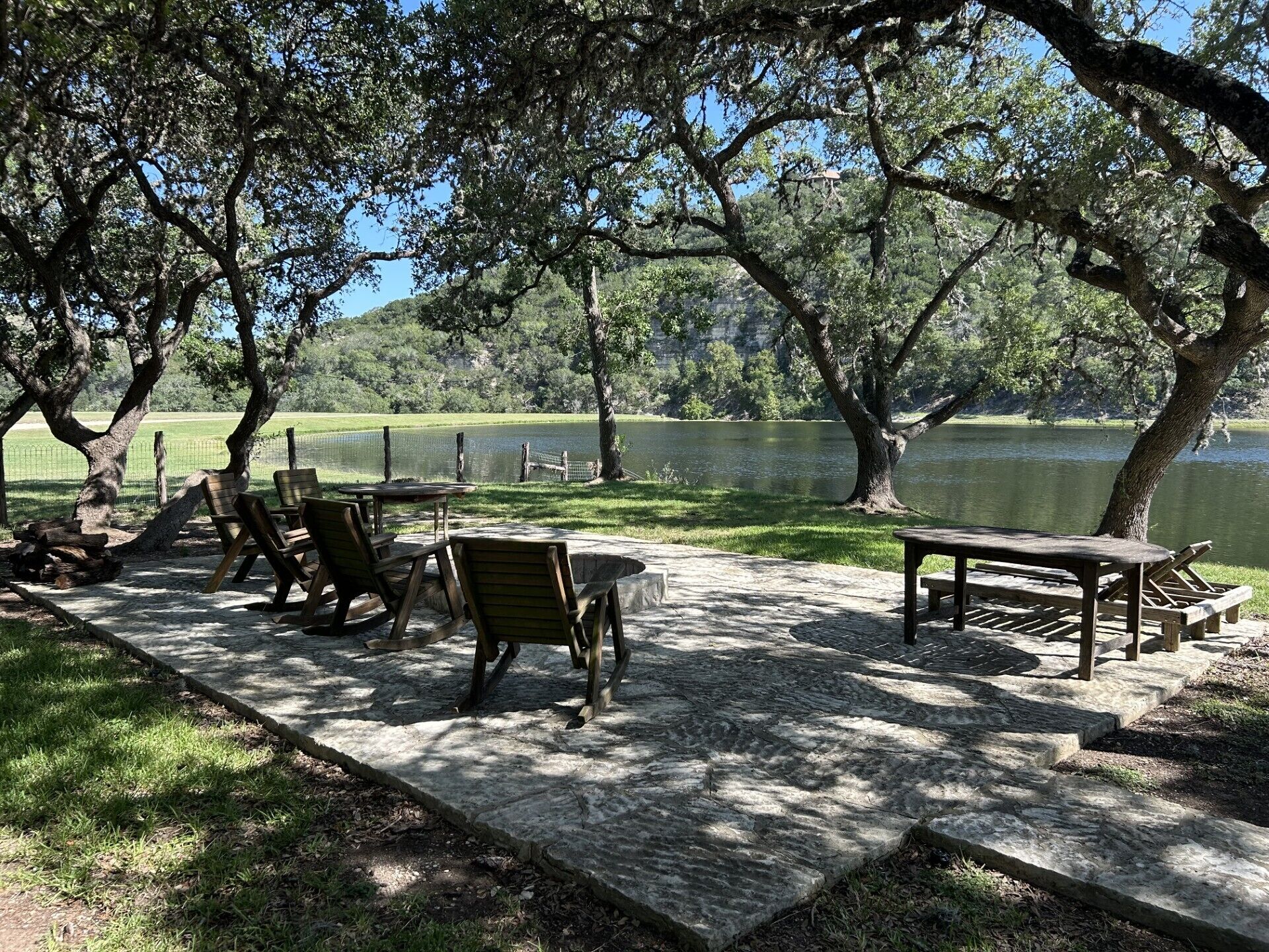 A picnic area with tables and chairs, a wooden bench, and a view of a lake surrounded by trees.