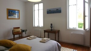 Ein Schlafzimmer mit einem Bett, zwei Fenstern, einem Sessel, einem Tisch und einem Bild an der Wand.