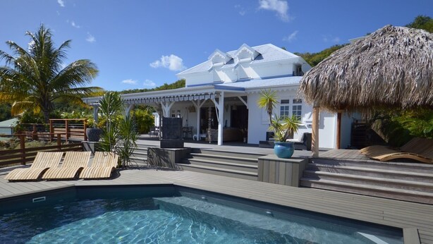 Ett poolområde med en klarblå pool, ett trädäck, solstolar och ett hus med ett halmtak.