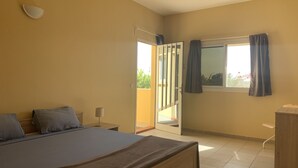 A bedroom with a bed, a nightstand, a lamp, a window with curtains, and a door leading to an outdoor area.