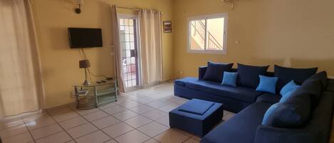 A living room with a blue sofa, a TV mounted on the wall, and a window with curtains.