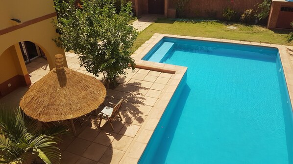 A swimming pool with a tiled deck, a thatched umbrella, and surrounding greenery. - Beautiful Riad Villa with Private Pool, Near Beach, Wifi (Saly)