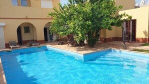 A swimming pool with a tree in the center, surrounded by lounge chairs and a patio area.