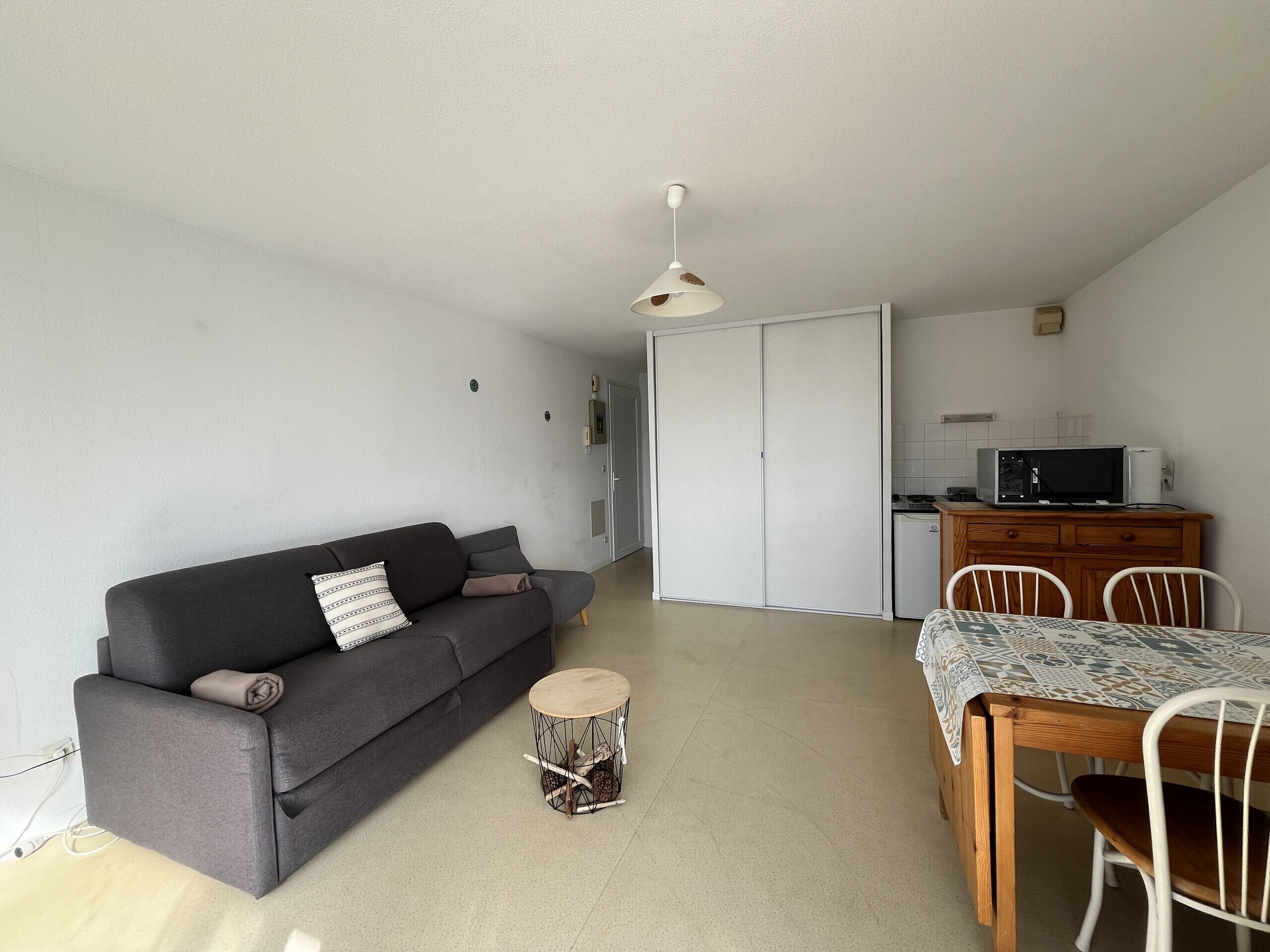 A compact living space with a grey sofa, a round wooden side table, a dining table with chairs, a kitchenette, and a sliding door closet.