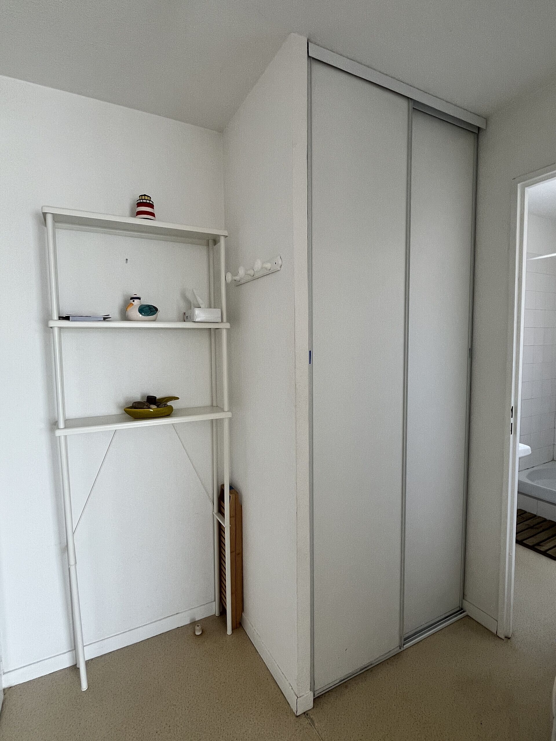 Ein Zimmer mit einem weißen Kleiderschrank, einem weißen Regalschrank mit verschiedenen Gegenständen und einer sichtbaren Tür zu einem anderen Raum.