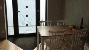 Un comedor con una mesa de madera y sillas blancas, una pequeña cocina con refrigerador, y un ventanal con vista a un edificio.