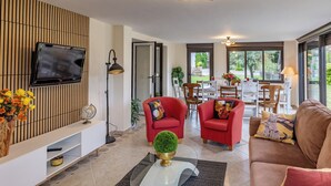 Un salon moderne doté d’un téléviseur à écran plat, de fauteuils rouges, d’une table basse en verre et offrant une vue sur l’extérieur grâce à de grandes fenêtres.