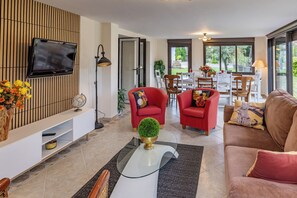 A modern living room with a flat-screen TV, red chairs, a glass coffee table, and a view of the outdoors through large windows. - Gite Les Bleuets - 12 pers. for get-togethers, near golf course (Saint-Geyrac)