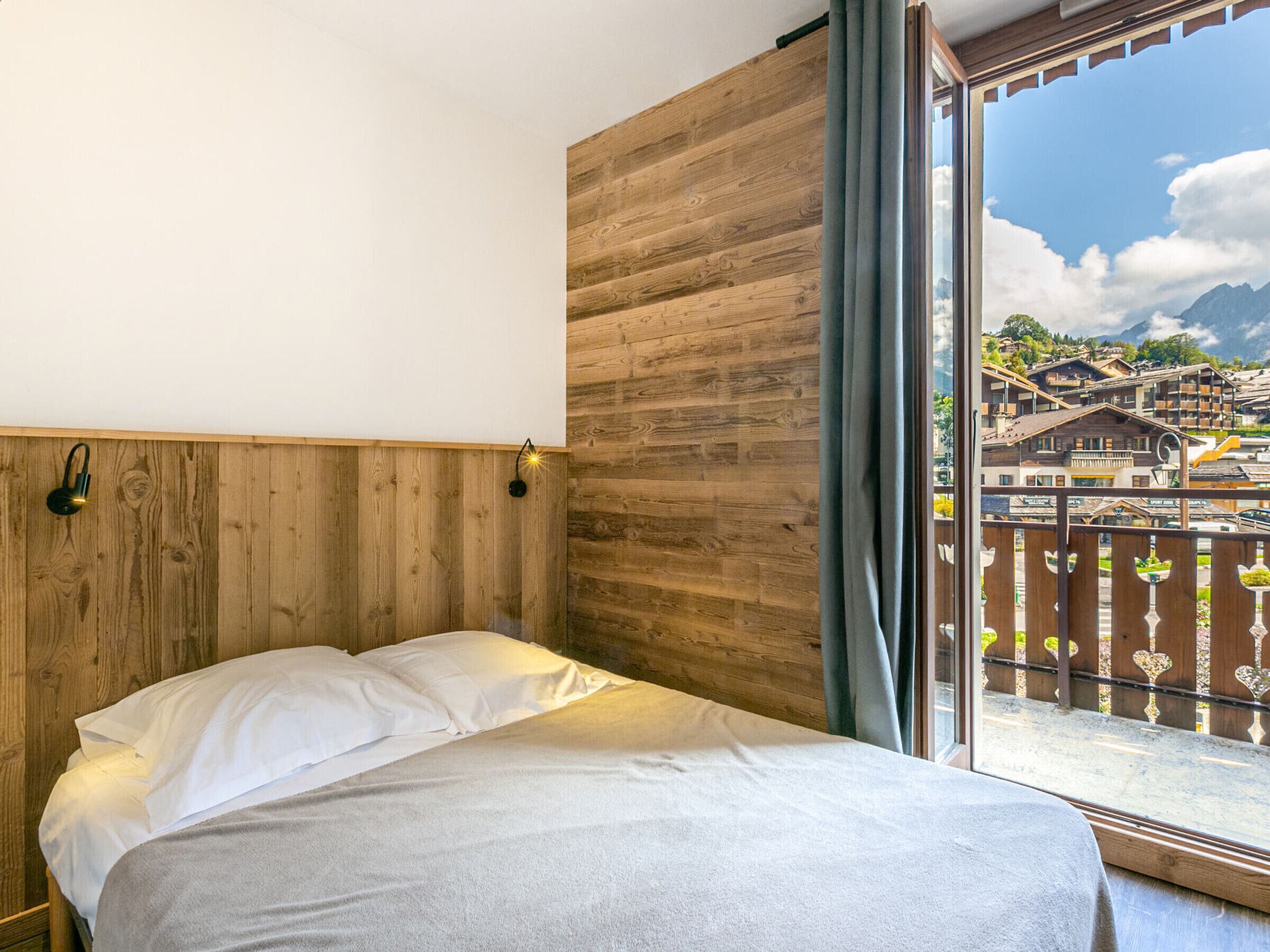 Ein Zimmer mit einem Bett, Holzwandverkleidung, einem Fenster mit Blick auf eine Bergstadt und einer Wandleuchte.