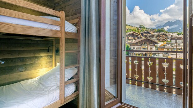 A room with bunk beds and a large window overlooking a town with buildings and mountains.
