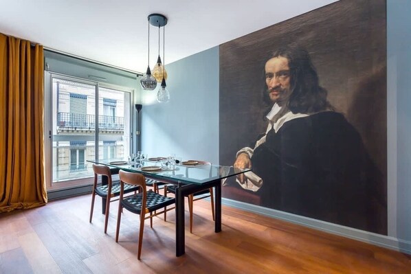 A dining area with a glass table, chairs, and a large portrait on the wall.
