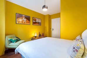 A room with a bed, a chair, a bedside table with a lamp, and two framed pictures on the wall.