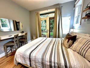 Un dormitorio con cama, escritorio y silla. Hay una ventana con cortinas y vistas a la vegetación.