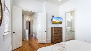 A bedroom with a bed, a dresser, a TV mounted on the wall, and a hallway leading to another room.
