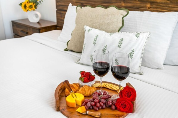 Una cama bien hecha con sábanas blancas y almohadas decorativas, una bandeja de madera con queso, uvas, fresas y un croissant, y dos copas de vino tinto.