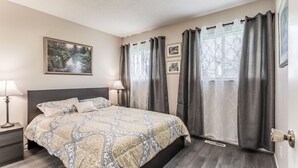Ein Schlafzimmer mit Bett, Nachttisch, Lampe, einem Fenster mit Vorhängen und einem gerahmten Bild an der Wand.