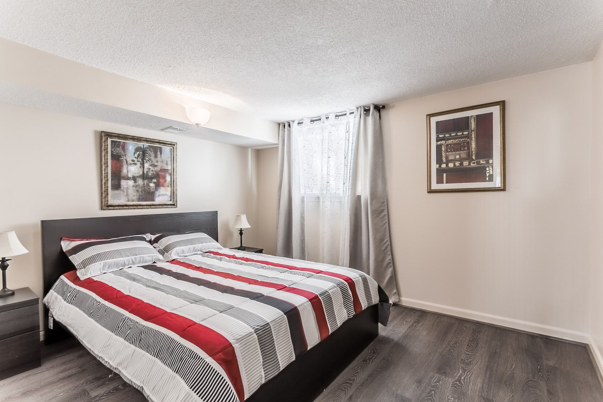 A hotel room with a bed, a nightstand, a lamp, a window with curtains, and two framed pictures on the wall.