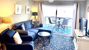 A living room with a sofa, coffee table, and a TV. There is a ceiling fan, a lamp, and a window with curtains.