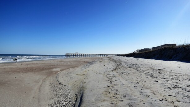 A beach with a wooden pier extending into the ocean, sandy terrain, and a clear sky.
