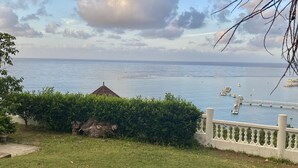 Zona verde con una valla blanca, una pequeña construcción y vista al mar con barcos.