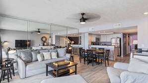 A modern open-plan living area with a sofa, coffee table, and dining table. A kitchen is visible in the background with a refrigerator and microwave. - EMERALD COAST PARADISE! 202 Destin Seafarer (Destin)