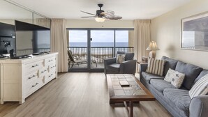 Een kamer met een bank, een salontafel, een televisie op een kast en een balkon met uitzicht op zee.