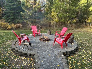 Et cirkulært opholdsområde med røde Adirondack-stole omkring et bålsted, omgivet af en stenkant og en grusgrund.
