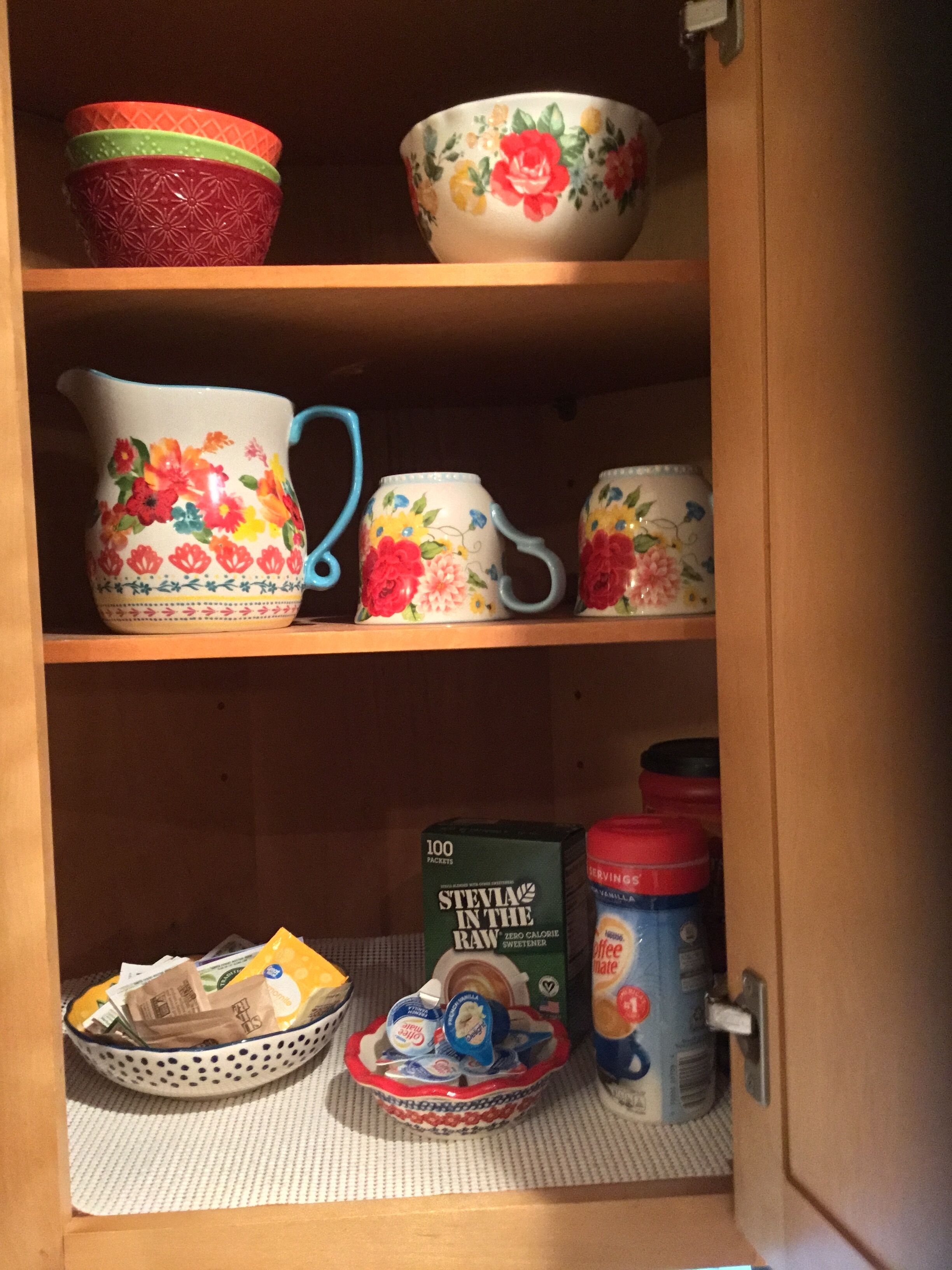 Un meuble de cuisine contenant divers objets, notamment un pichet à motifs floraux, des tasses, des bols et un récipient rempli de portions individuelles de yaourt.