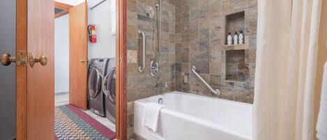 Un baño moderno con pared de piedra, una bañera, ducha con barras de apoyo y zona de lavandería.