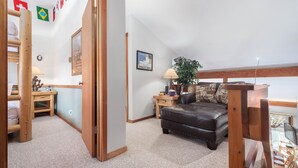 Un salon chaleureux avec un canapé en cuir marron, une table basse en bois et une petite table d’appoint avec une lampe. Une échelle en bois mène à une mezzanine dotée d’une grande fenêtre.