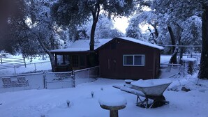 A snow-covered outdoor area with a wooden bench and a wheelbarrow, a red building with a window, and a fence. - The High Country Hobo Preserve: Rustic Cabin (Descanso)