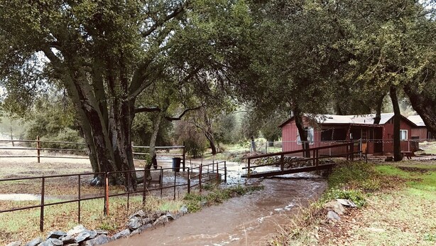 A rural scene with a muddy stream, a wooden bridge, and a red building with a porch. - The High Country Hobo Preserve: Rustic Cabin (Descanso)