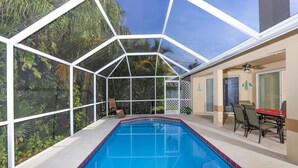 Une piscine entourée d’une paroi de verre, avec des chaises et une table, et offrant une vue sur la verdure environnante.