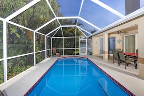 A pool with a glass enclosure, surrounded by chairs and a table, with a view of greenery outside. - Beautiful Pool Home Close To Many Beaches (Venice)